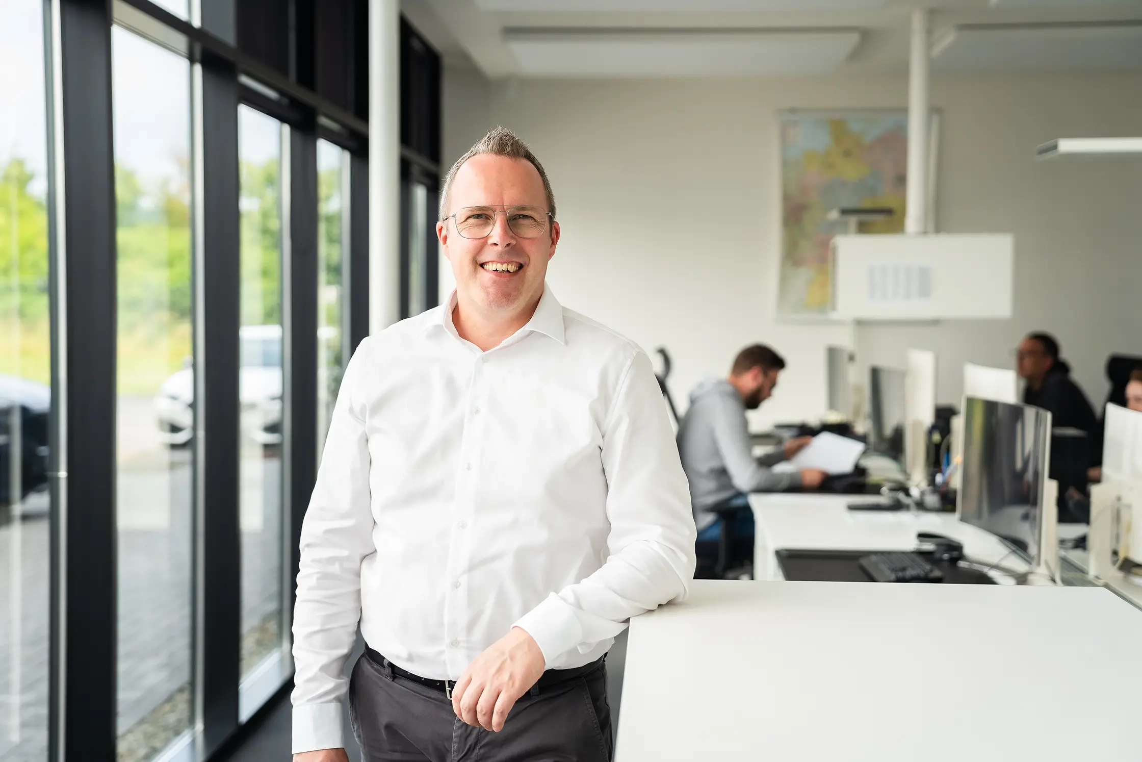 Geschäftsführer Michael Jost steht in einem modernen Büro mit großen Fenstern. Er trägt ein weißes Hemd und schwarze Hose. Im Hintergrund sind Mitarbeiter an Computern zu sehen.