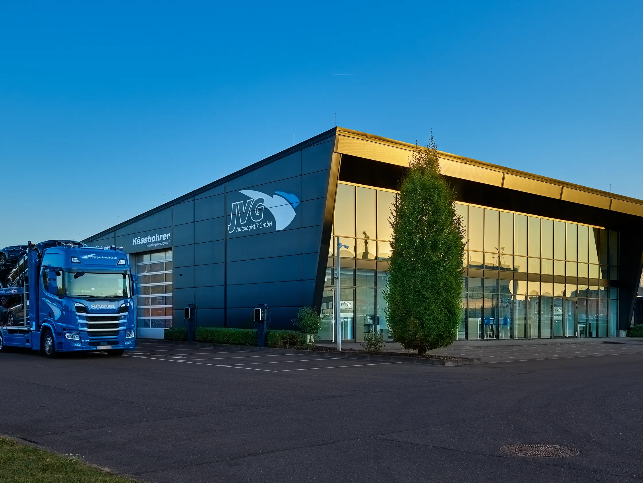 Das JVG Firmengebäude mit reflektierendem Sonnenuntergang auf der Fassade. Ein blauer Lkw steht auf dem Parkplatz.