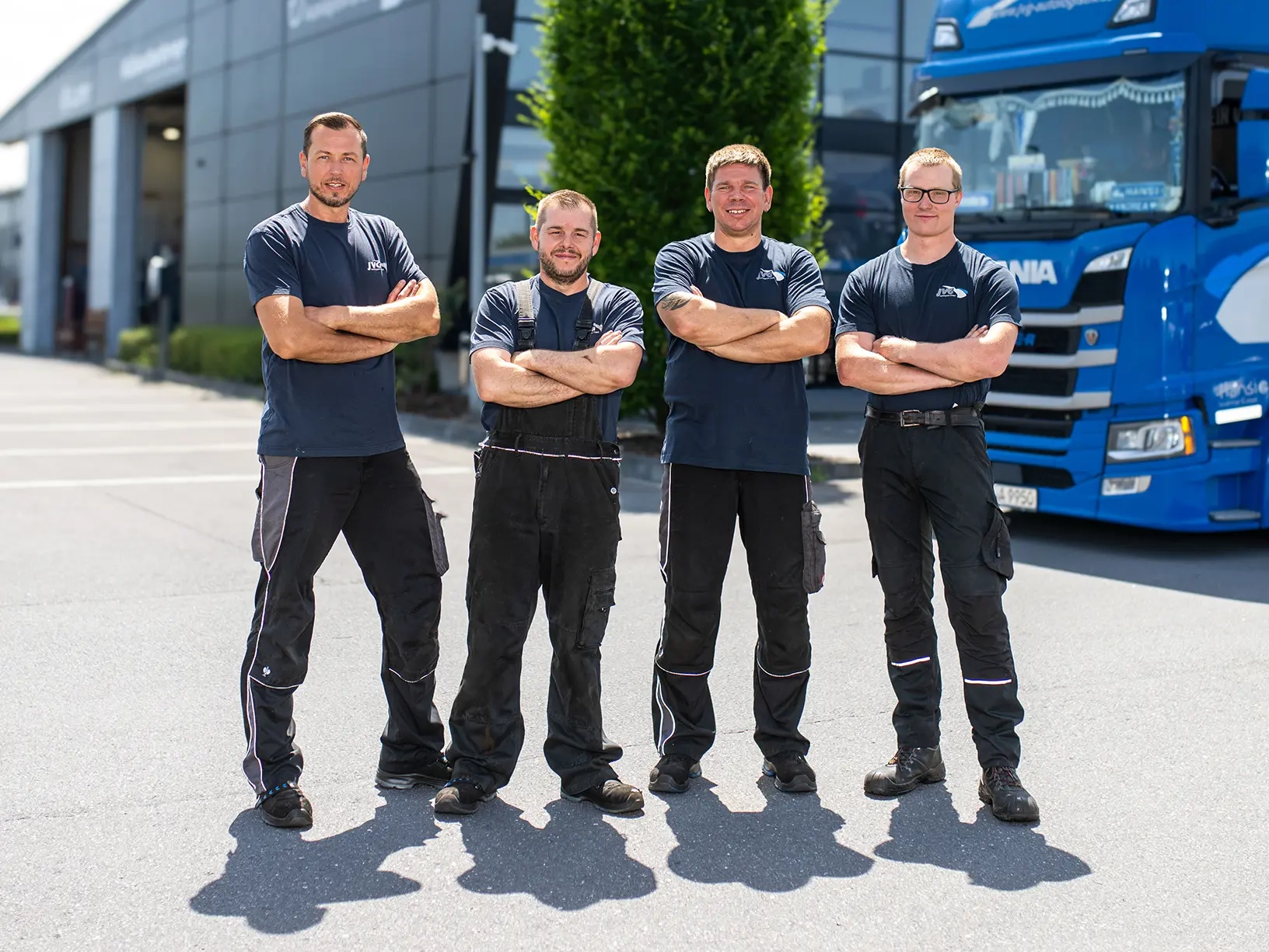 Vier Arbeiter in blauen T-Shirts und schwarzen Hosen stehen mit verschränkten Armen auf einem Parkplatz vor einem Lkw.