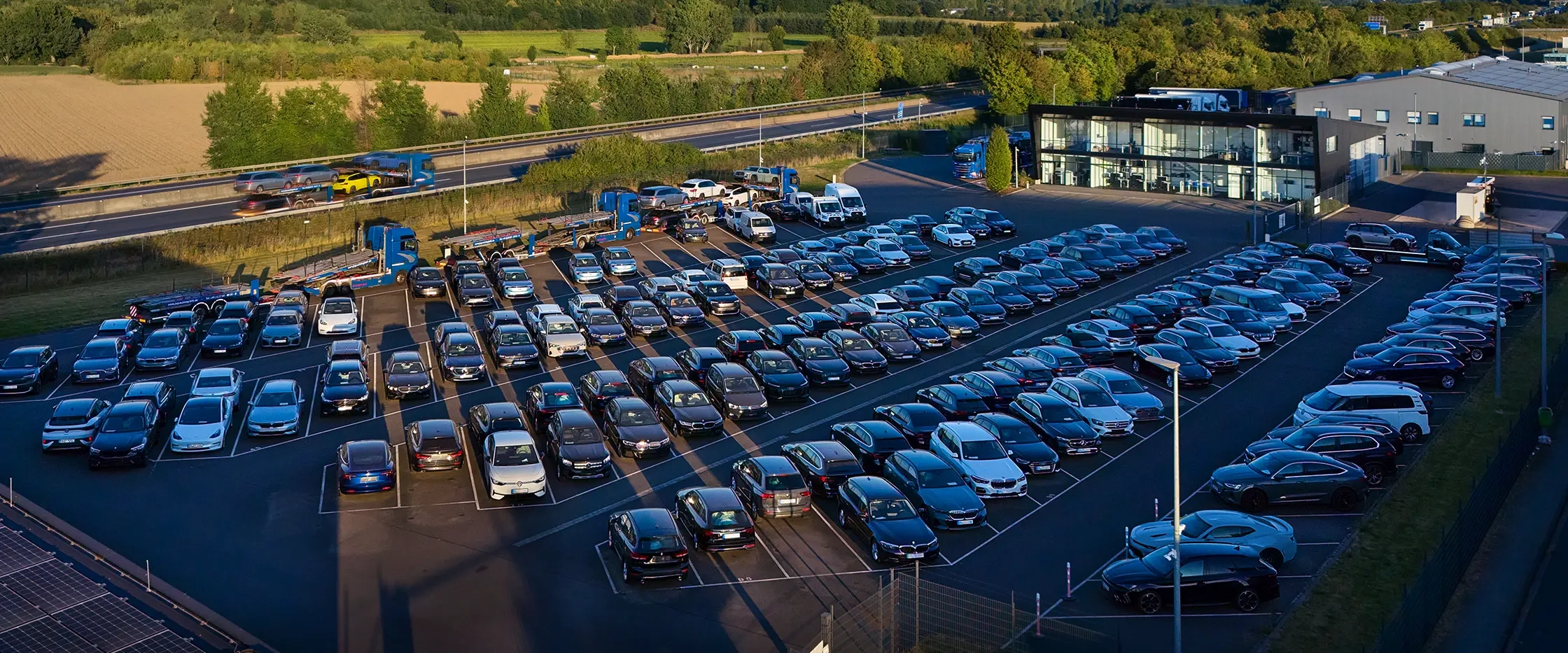 Luftaufnahme des JVG Firmengebäudes mit einem großen Parkplatz, der mit vielen Autos belegt ist. Im Hintergrund verläuft eine Straße mit Fahrzeugen.