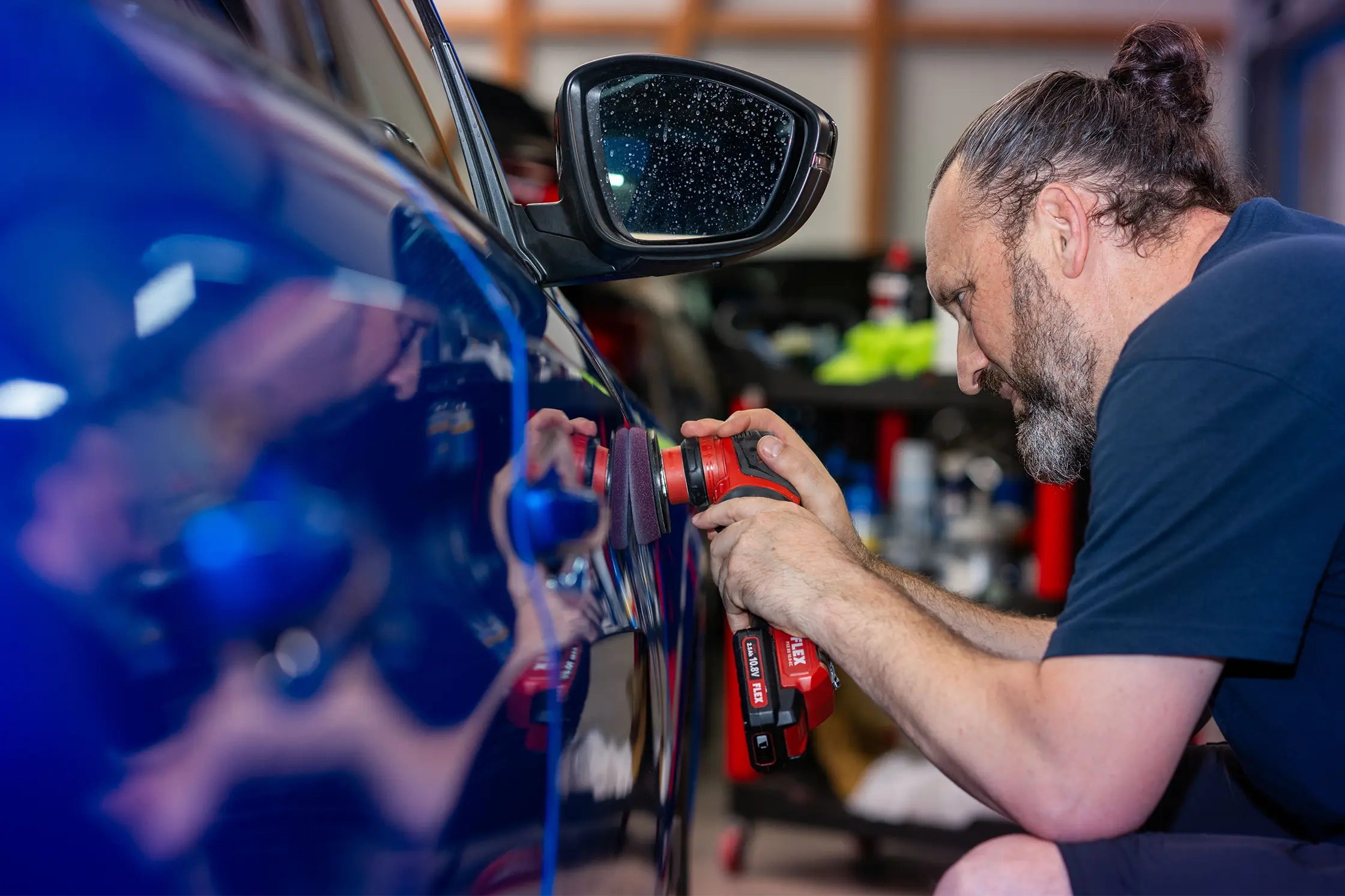 Ein Mitarbeiter von JVG poliert ein blaues Auto mit einer Poliermaschine. Im Hintergrund sind Werkzeuge und Ausstattungen sichtbar.
