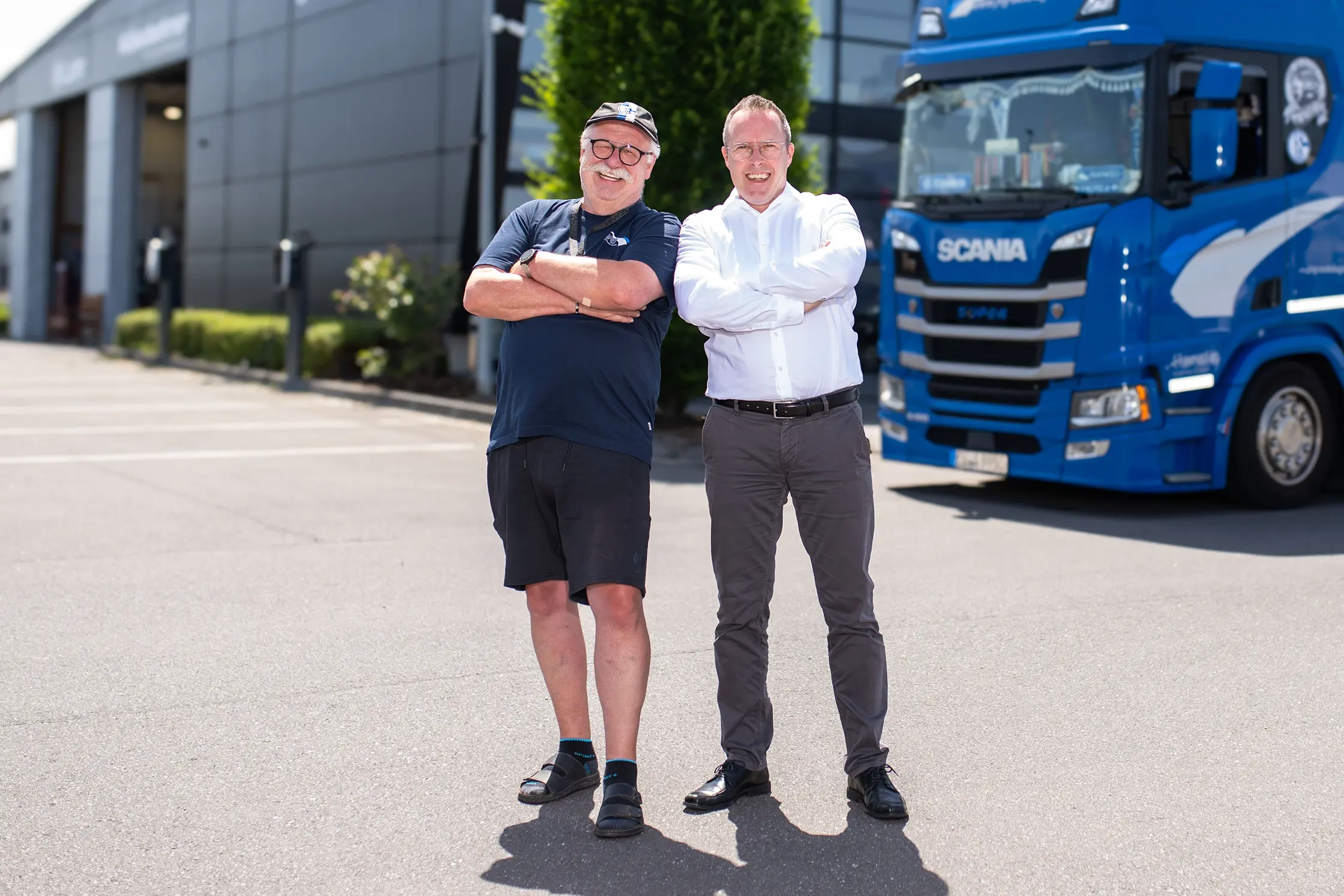 LKW Fahrer steht links, trägt dunkle Kleidung und Sandalen. Geschäftsführer Michael Jost steht rechts in einem weißen Hemd und grauen Hosen. Im Hintergrund ein blauer Scania Lkw.