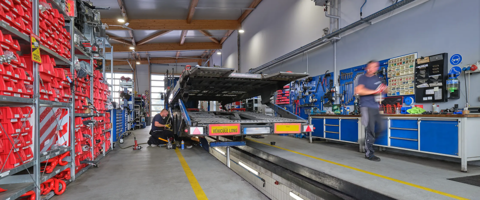 JVG Mitarbeiter arbeiten in der Werkstatt, ein Mitarbeiter arbeitet an einem Lkw mit offener Ladefläche, Werkzeugregale an den Wänden, rote Werkkästen sind sichtbar.