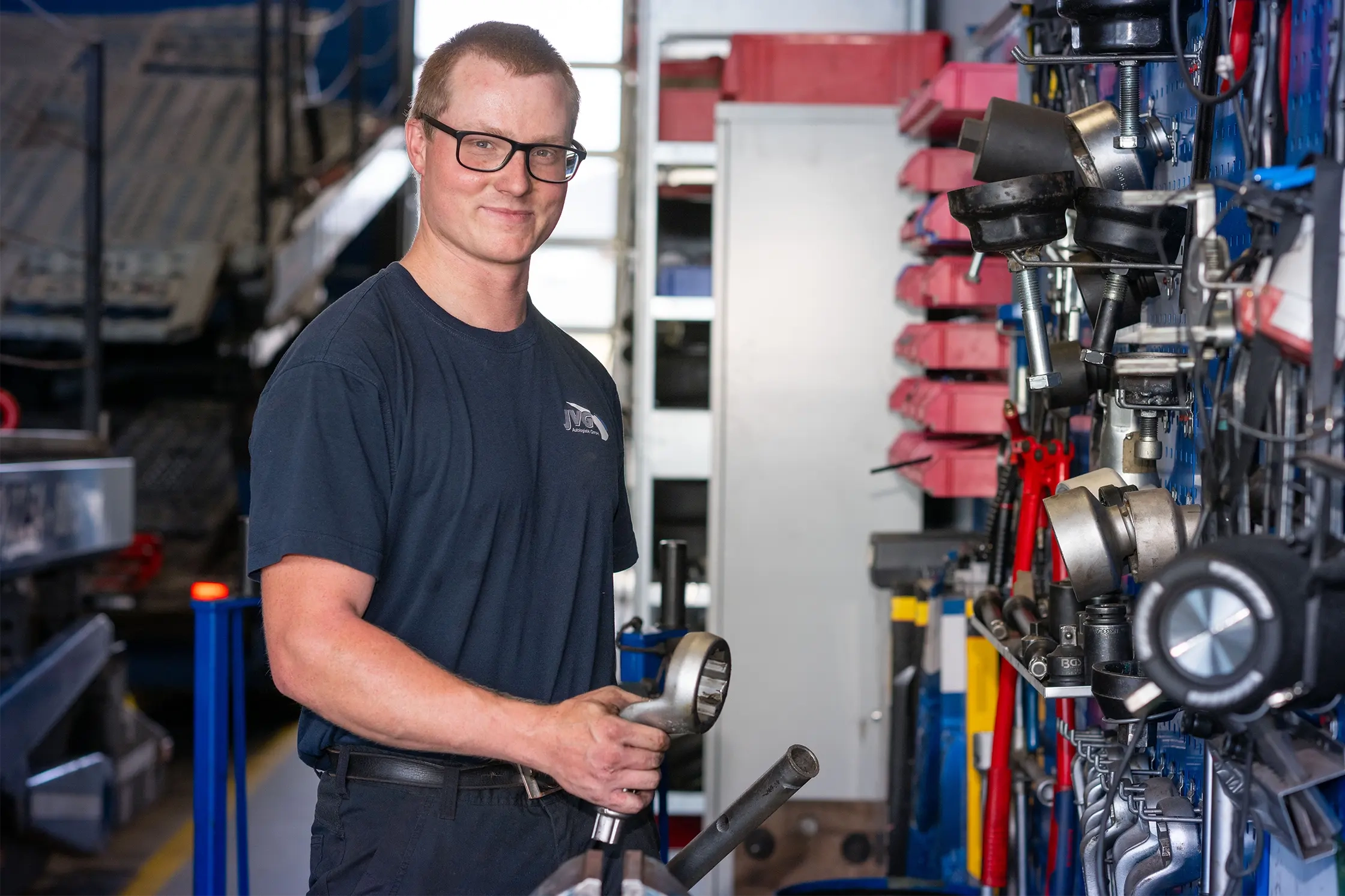 Mitarbeiter von JVG in Arbeitskleidung, hält ein Metallrohr in der Hand, steht vor einem Werkzeugregal mit verschiedenen Werkzeugen.
